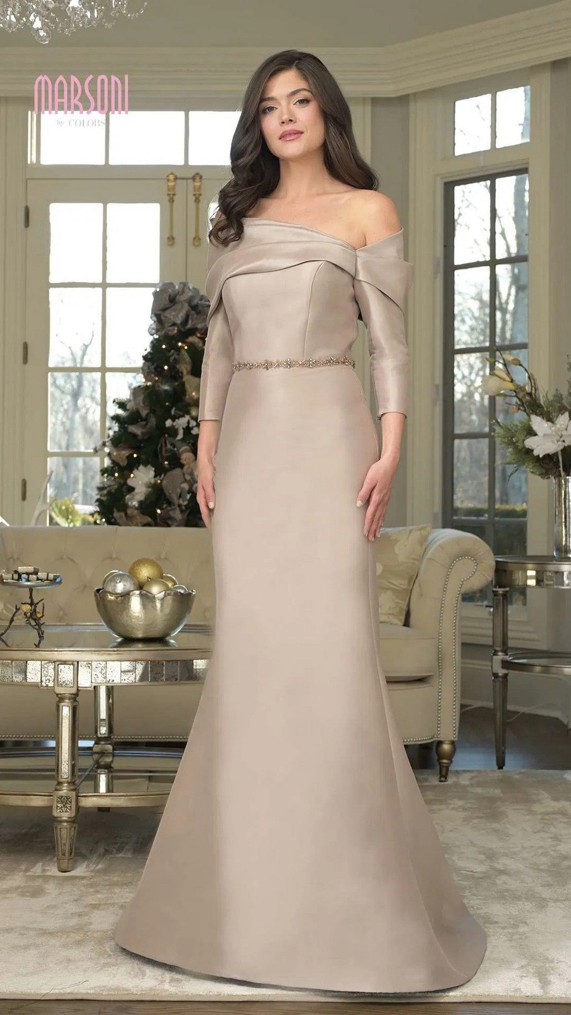A woman in the Marsoni MV1375 long formal dress in taupe with an off-shoulder neckline stands in a living room decorated with a Christmas tree and holiday ornaments.
